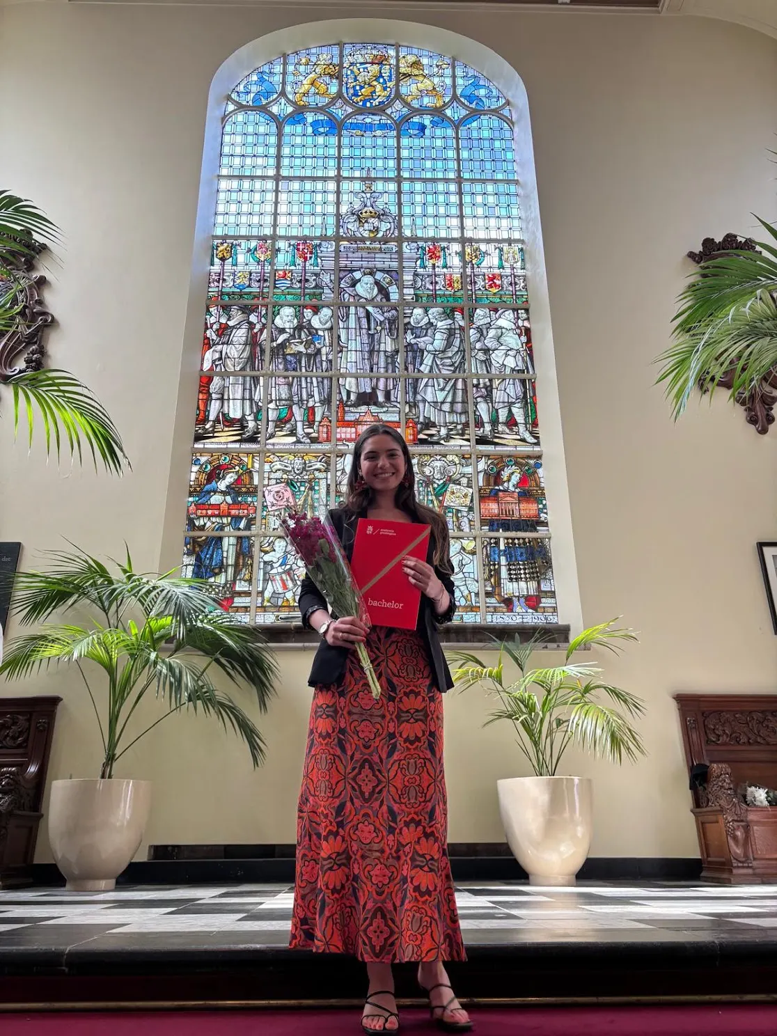 Raquel holding flowers and her diploma in a hall with a large stained-glass window