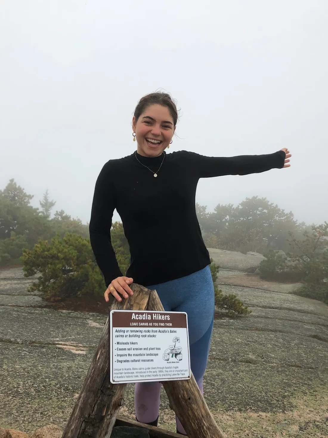 Raquel smiling next to an Acadia hikers sign on a foggy granite summit in Maine
