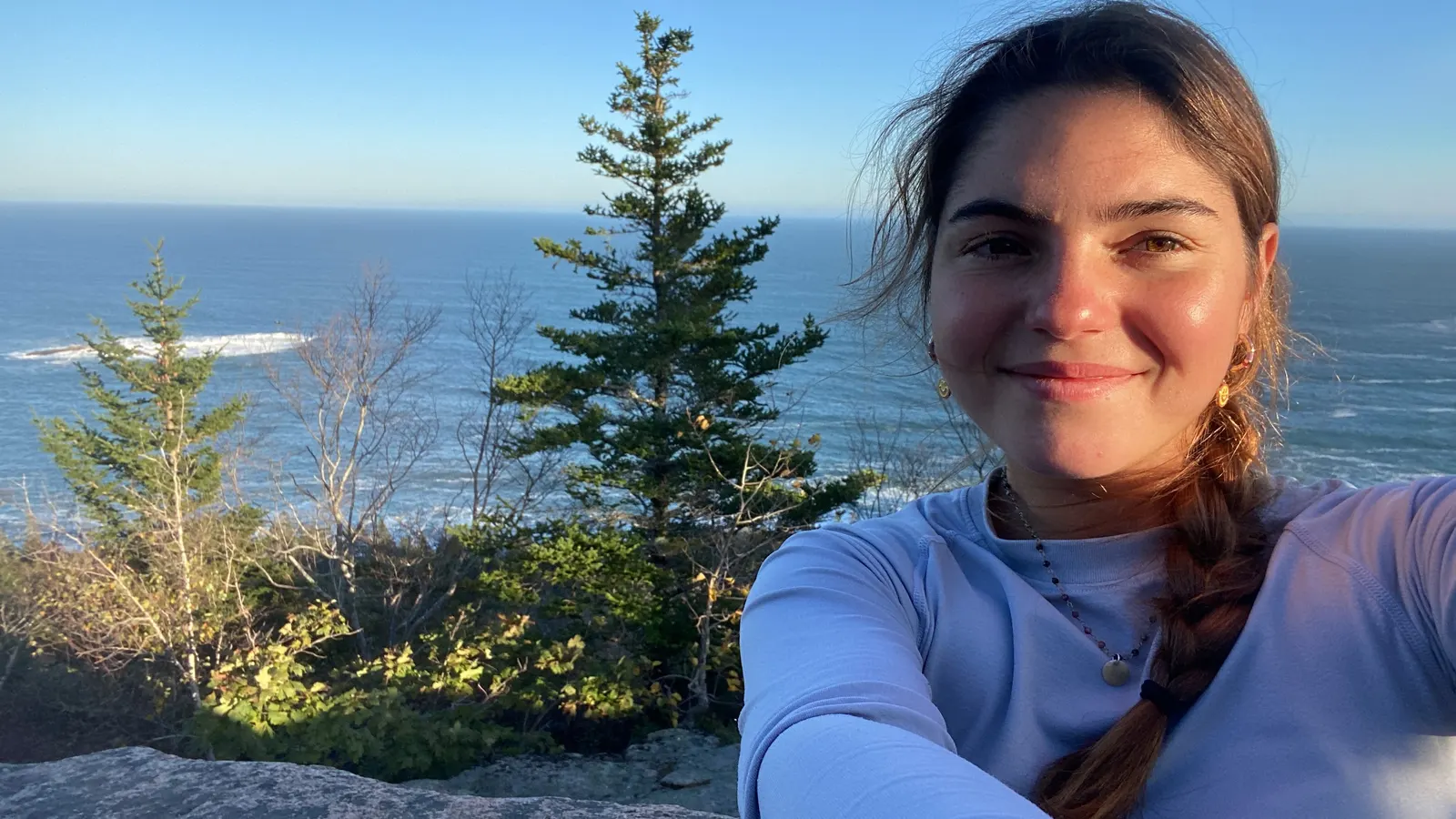 Selfie of Raquel on the rocky coast of Maine with pine trees and the ocean behind